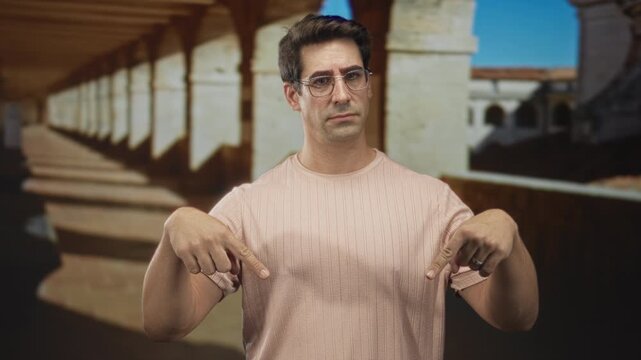 Man wearing glasses points both fingers downward at his pink t shirt in a building corridor with stone arches and columns; skepticism instruction.