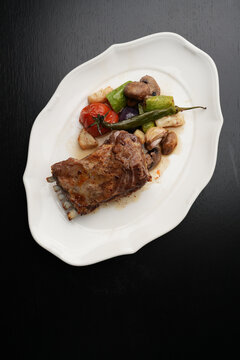 Roasted Lamb Rack with Grilled Vegetables and Chili Pepper. Baked lamb ribs served with grilled tomatoes, zucchini, mushrooms, and a whole green chili pepper. Gourmet meat dish on a white plate