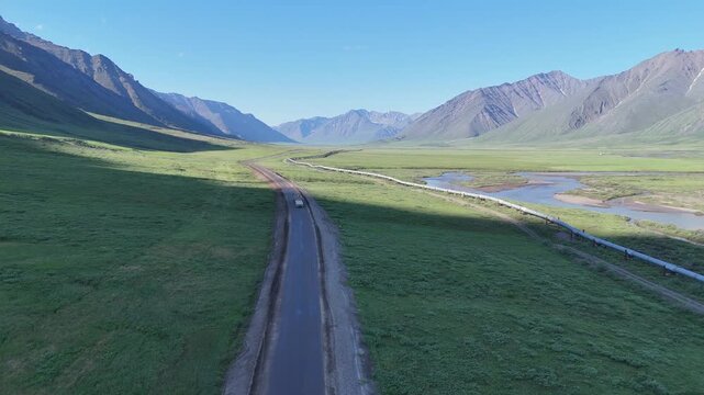Aerial view of a white truck driving on the famous dalton highway, crossing a vast green valley in alaska with the trans alaska pipeline running alongside the road and a river
