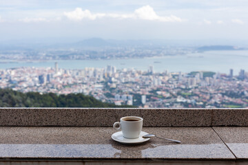 Obraz premium Hot coffee cup on balcony overlooking Penang Hill. Morning beverage with cityscape view in Malaysia. Scenic urban landscape from elevated cafe terrace.