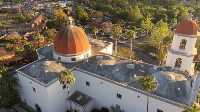 Aerial drone shot gliding above Mission Basilica San Juan Capistrano during golden hour, highlighting the copper dome, bell tower, palm trees, and surrounding streets in historic San Juan Capistrano,