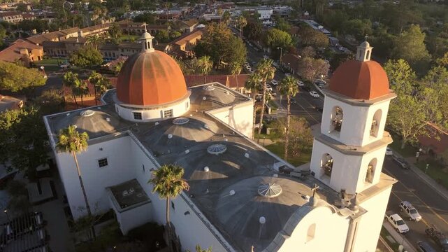 Aerial push-forward drone shot of Mission Basilica San Juan Capistrano in warm golden hour light, with the historic San Juan Capistrano Mission visible in the background, palm trees and downtown