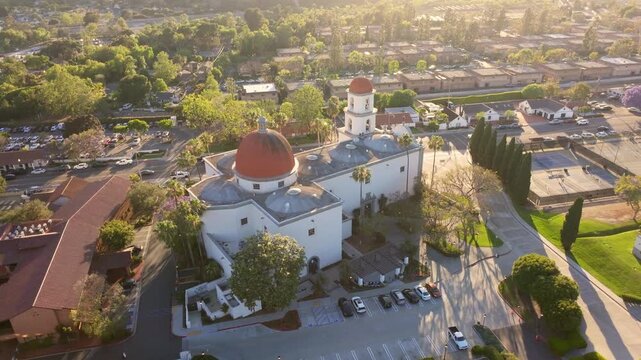 Wide aerial drone shot of Mission Basilica San Juan Capistrano during golden hour, showing the church dome, bell tower, parking area, and surrounding neighborhood in historic San Juan Capistrano