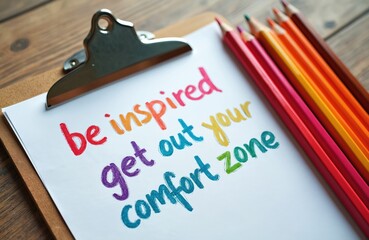 Obraz premium Colorful paper on clipboard with text be inspired get out your comfort zone and colored pencils. Message promotes change, growth, and new experiences.