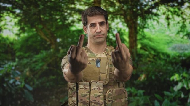 Man in camouflage tactical vest shows both middle fingers, dog tags visible, standing in forest; defiance protest.