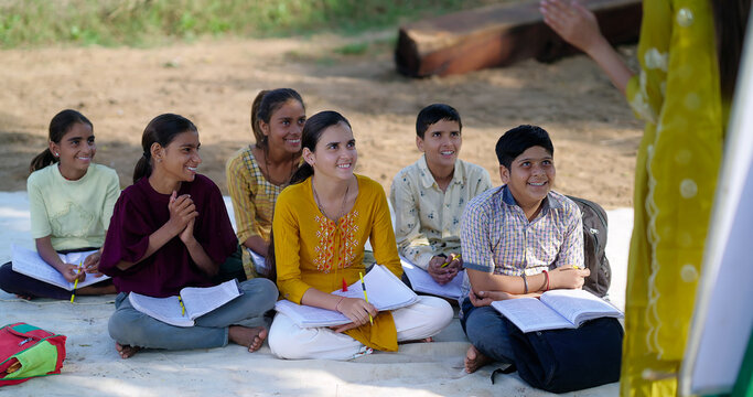 Indian adult female lady talk speak teach to villager group day under tree girl boy sitting on mat do study look board learn at open class field rural area place