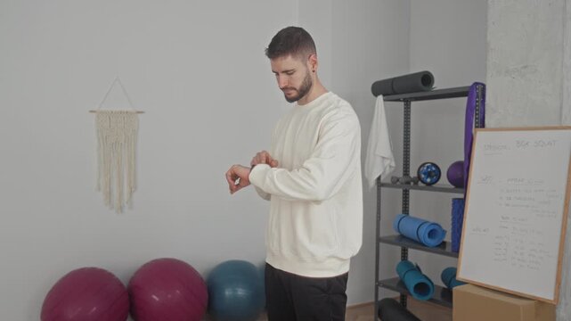Man checks watch while standing by yoga balls, rolled mats and a whiteboard in a studio; focus preparation.