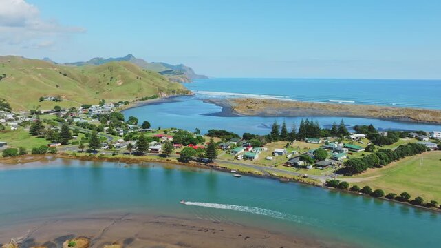 A boat speeds down the turquoise waters of the River in Marokopa, Waikato, New Zealand, passing by lush green hills and a small coastal town.