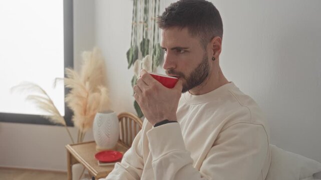 Young man sipping from red cup on bed in bedroom, holding mug to lips and looking contemplative; calm reflection.