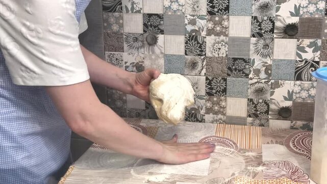 Female hands kneading raw dough on a patterned table, adding flour, traditional baking, home kitchen lifestyle.