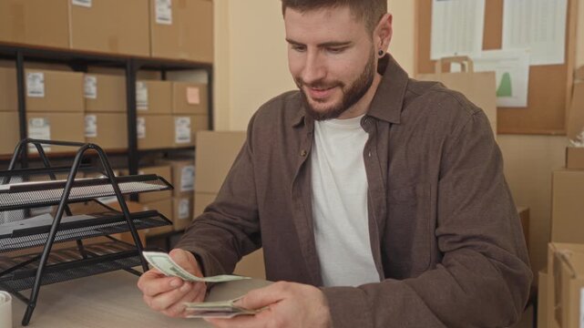 Man counts cash with hands at a warehouse building desk surrounded by boxes and packing materials; small business focus.