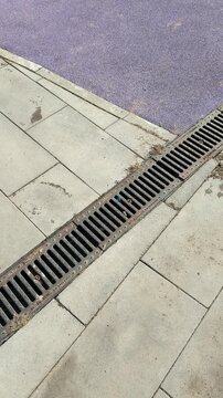 Iron drainage grate on sports pavement, rusty metal plate set in linear channel between concrete pavers, dried leaves and grit.