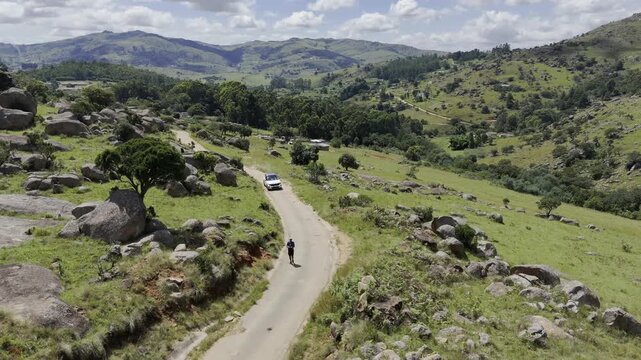 Drone orbits right over trail as car approaches hiker on a sunny day at Sibebe Rock near Mbabane, Eswatini