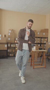 Man holding small parcel and checking smartphone while walking among stacked boxes in building; focused logistics.