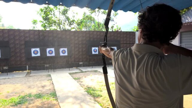 Young man aiming bow at target range outdoors from back view, archery practice session, focus and precision concept, active leisure sport, skill training activity.