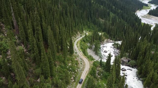 Aerial high angle shot of an off-road SUV driving on a winding dirt road along a raging mountain river in a deep forest gorge, Kyrgyzstan, Karakol.
