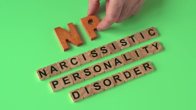 A hand arranging the abbreviation NPD with wooden letters.  Its explanation: Narcissistic Personality Disorder, formed with wooden letter tiles. Light green background. 