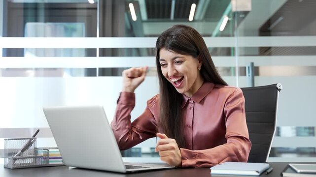 Happy businesswoman sitting at desk in office, reading good news on laptop and celebrating success. Excited female employee feeling joy and satisfaction after receiving positive message in workplace.