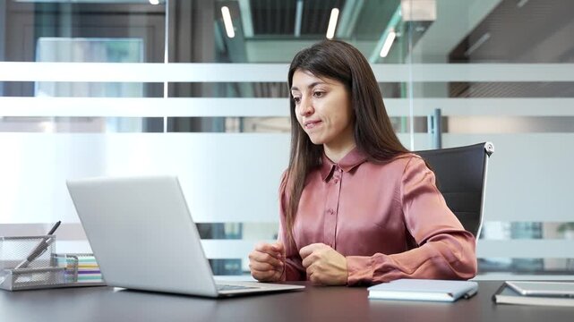 Businesswoman arguing during video call in office, looking at laptop screen. Upset female expressing frustration and stress, involved in a conflict while communicating online in corporate workplace.