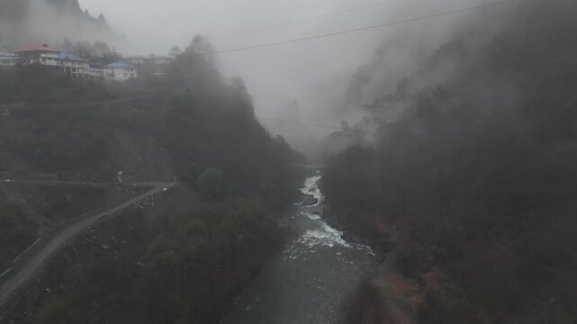 Aerial drone footage captures a misty mountain river flowing through a deep valley with fog rolling between forested hillsides and a small village with colorful rooftops perched on the steep slopes ab