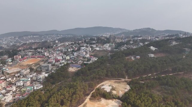 Drone footage reveals a sprawling hillside town with colorful multi-story buildings nestled among lush green pine forests. Mountains rise in the misty background as winding roads cut through the terra