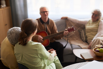 Senior man playing guitar for senior residents and female caregiver in nursing home, supporting...