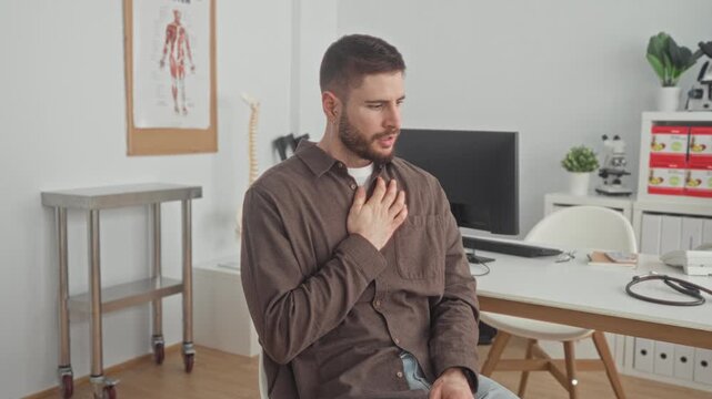 Man clutching chest seated at a desk with stethoscope in a medical building, coughing and wincing; anxiety illness.