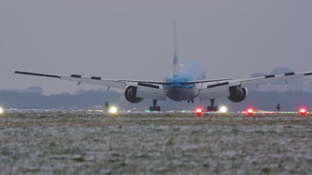 Rear view of commercial aircraft touching down on snow-dusted runway at dusk, spoiler deployed, colorful runway light and city skyline in background
