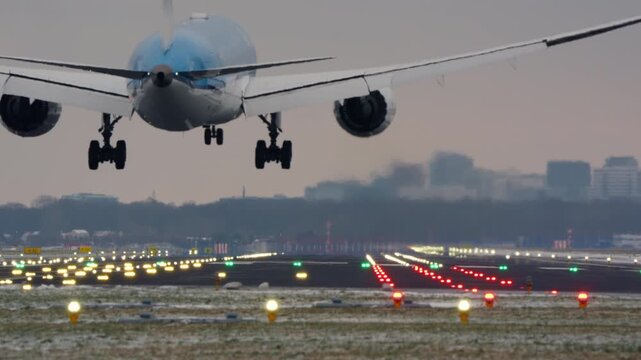 Front underside view of commercial aircraft on final approach with landing gear deployed over illuminated runway threshold in winter, city buildings in background