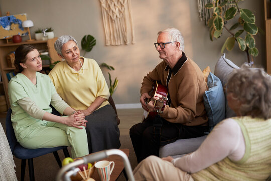 Senior man playing guitar for senior women and young adult caregiver in nursing home, supporting social engagement