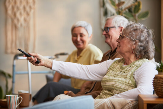 Senior Caucasian woman using remote control while sitting with senior friends in nursing home, representing assisted living leisure