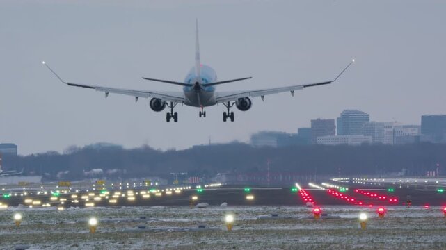 Rear view of commercial aircraft on final approach with landing gear extended over runway threshold light, winter city skyline and buildings in background