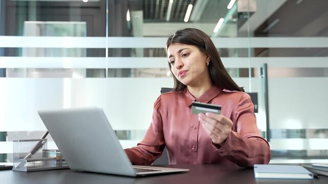 Upset businesswoman sitting in office, shocked after detecting credit card problem on computer. Worried female employee realizing fraud or payment error, stress and disbelief in corporate workplace.