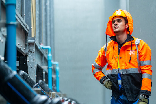 Industrial worker in raincoat inspecting pipeline system in factory during heavy rain for maintenance and engineering technical inspection industrial equipment repair troubleshoot safety work process