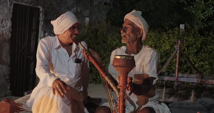 Indian old aged man wear white kurta pajama sitting on woven cot bed enjoy desi life at rural area. Happy two male group talk speak share gossip do hookah hold pipe spend night time village place