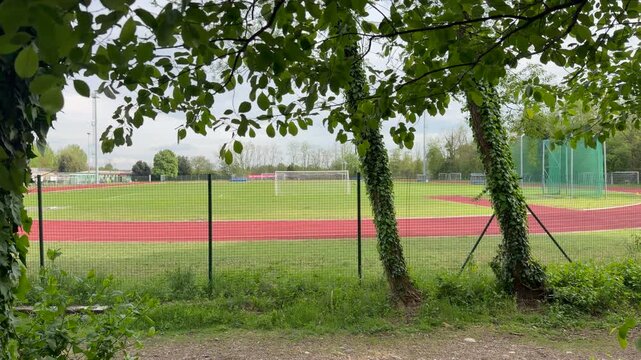 View of sports field and running track framed by trees and fence, outdoor athletic area in natural setting, cloudy sky and green surroundings, recreation and leisure concept.