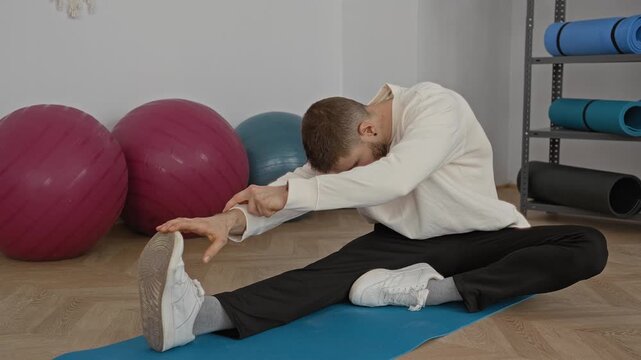 Man seated on blue yoga mat reaches to touch shoe while stretching beside exercise balls in studio; calm focus.