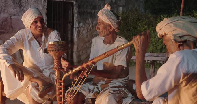 Indian old aged man wear white kurta pajama sitting on woven cot bed enjoy desi life at rural area. Happy three male group talk speak share gossip do hookah hold pipe spend night time village place