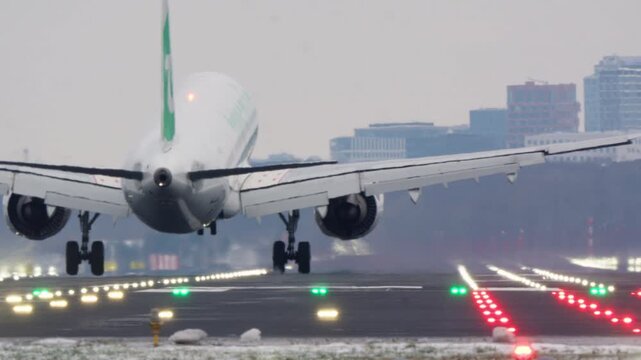 Close-up underside view of commercial aircraft on short final with landing gear down, runway approach light in foreground, winter haze and city in background
