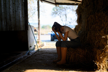 Exhausted female farm worker sitting on hay bale in barn feeling stressed and overwhelmed