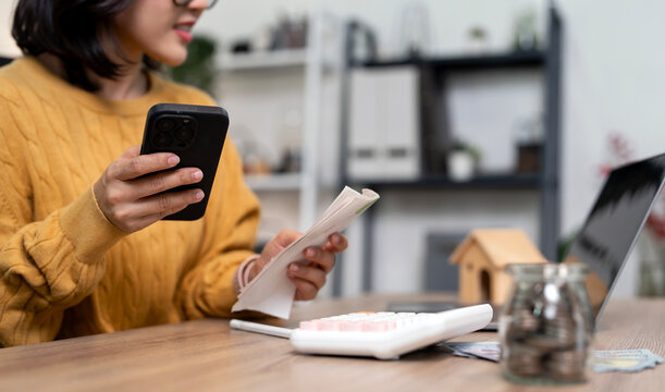 Asian woman using smartphone for mobile banking and managing household expenses in a home office.