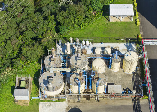 Aerial view of a beverage factory corner with large storage tanks, blue pipelines, processing equipment, and surrounding green trees on a sunny day.