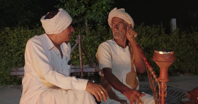 Indian old aged man wear white kurta pajama sitting on woven cot bed enjoy desi life at rural area. Happy two male group talk speak share gossip do hookah hold pipe spend night time village place