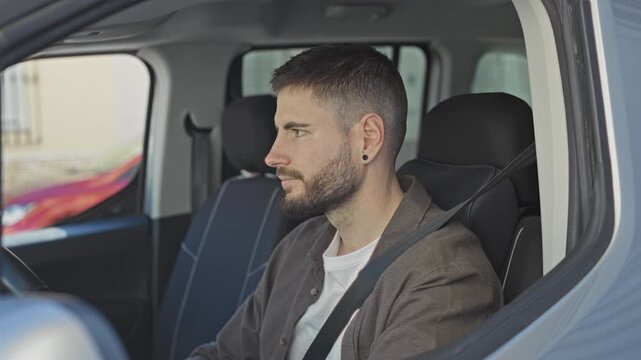 Man in car wearing seatbelt, touching ear and chin while looking out window from driver's seat on street; contemplation.