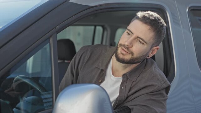 Man in car reaches hand out through open window toward side mirror while leaning from driver seat on street; request help.