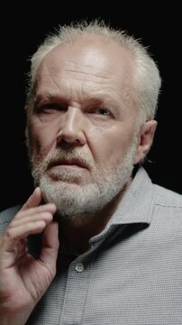 Senior Caucasian man touching face and looking down. Male pressing hand to cheek and showing concern. Thinking about problem and feeling tired. Standing in dark studio background.
