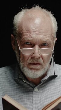 Caucasian man wearing glasses closing old book and raising dust. Male releasing particles into air and watching them. Reacting to floating dust with focused look. Standing in dark studio background.
