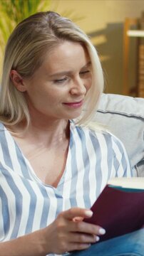 Caucasian woman sitting on sofa reading book and smiling. Female turning page with hand and focusing on text. Enjoying quiet moment at home. Resting in cozy living room. Vertical orientation.