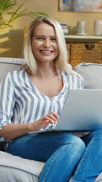 Smiling Caucasian woman sitting on sofa and using laptop. Positively looking at camera. Female typing on keyboard and reading screen. Working online and enjoying time in cozy room.