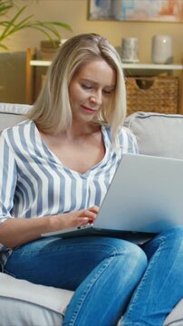 Caucasian woman using laptop while sitting on sofa and working remotely. Female typing on keyboard and focusing on tasks. Managing freelance job from home. Spending time in comfortable room.